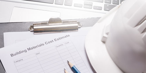 Clip board of a materials cost estimate sheet laying on top of a laptop with a hard hat nearby